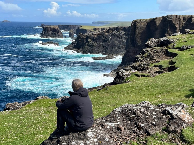 Person sits on a rock overlooking Shetland's rugged coastline with crashing waves, part of Shetland Wildlife.