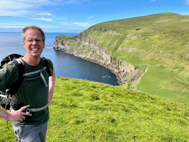 Hiker on Foula island, Shetland, with stunning green cliffs and ocean views.