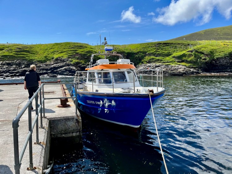 Blue boat "Silver Swan" docked in Foula, Shetland, with green hills and a person on the pier.