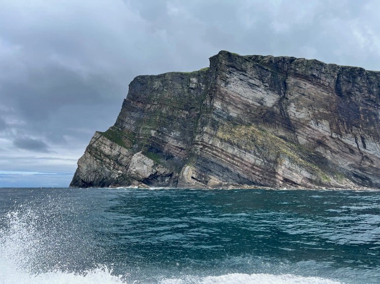Foula island's layered cliffs rise from the sea; Shetland wildlife habitat under a cloudy sky.