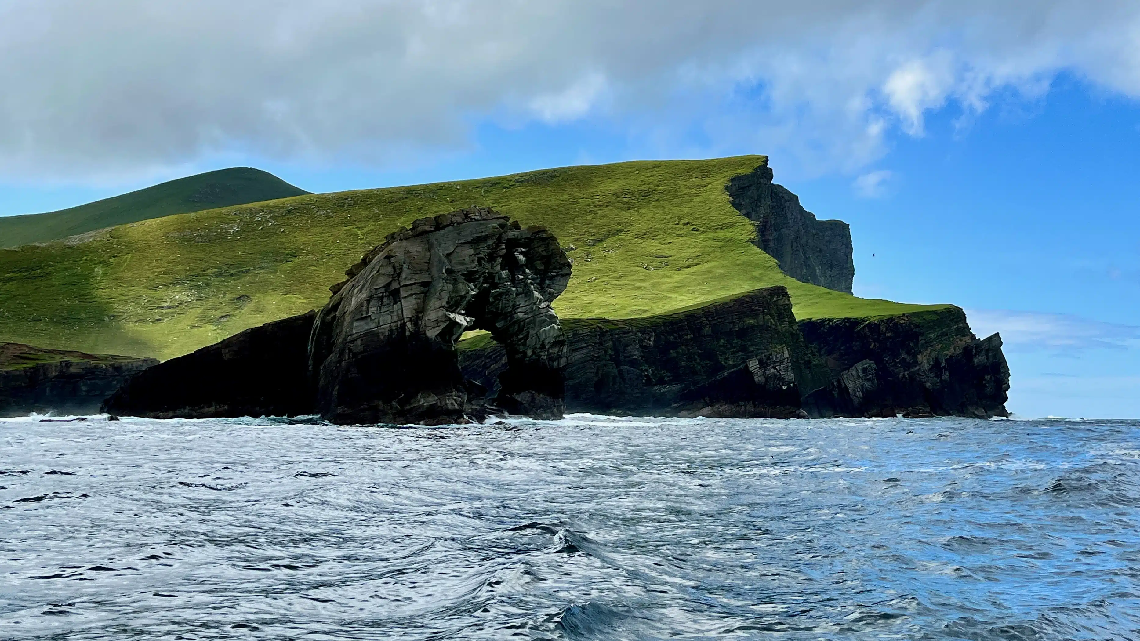 Foula island sea arch, Shetland. Rugged coastal landscape with green hills and dark cliffs.