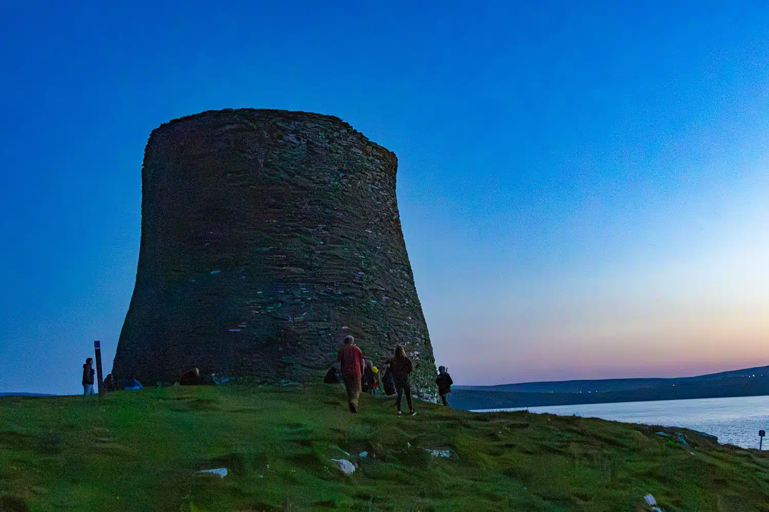Mousa Broch, an Iron Age round tower in Shetland, with people exploring the green hilltop at twilight.