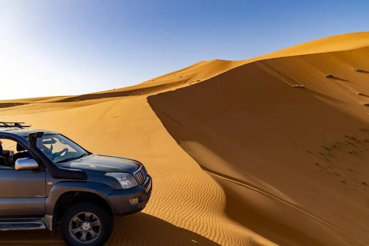 SUV driving on sand dunes in Morocco. Blue sky above desert landscape.