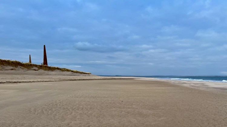 Northumbria beach with dunes and two brick towers under a cloudy sky.