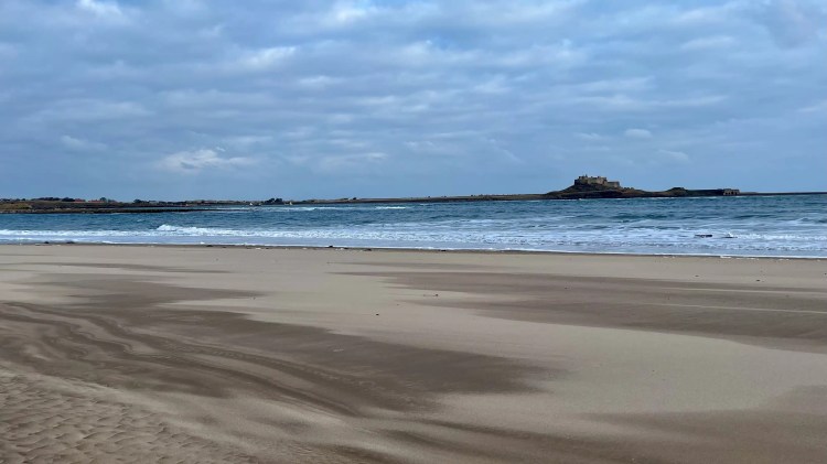 Northumberland beach view with waves and Lindisfarne Castle under a cloudy sky. Birding weekend location.